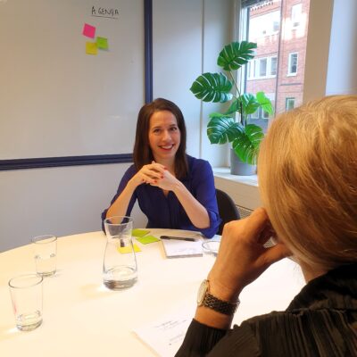 Frida Malmgren sitter vid ett runt bord mitt emot en person som hon handleder. På bordet finns papper och vattenglas. På väggen bakom henne finns en whiteboard.